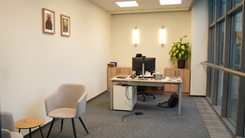 Jasne, nowoczesne biuro w siedzibie Bonifraterskiego Centrum Medycznego. Po prawej stronie ciąg dużych okien z widokiem na sąsiedni budynek. W głębi stoi biurko z dwoma monitorami, klawiaturą, dokumentami i kablami zwisającymi na podłogę. Za biurkiem ustawione są niskie szafki, na jednej z nich duża doniczka z zieloną rośliną. Po lewej stronie zdjęcia widać dwa beżowe fotele i mały okrągły stolik, a nad nimi na ścianie wiszą dwa portrety zakonników w ramkach