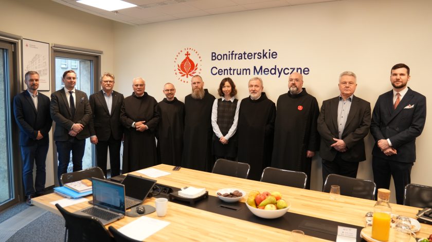 Grupa jedenastu osób – braci zakonnych w czarnych habitach oraz przedstawicieli zarządu w eleganckich garniturach – stoi w szeregu na tle ściany z napisem „Bonifraterskie Centrum Medyczne”. Przed nimi widoczny jest stół z laptopami, dokumentami i poczęstunkiem