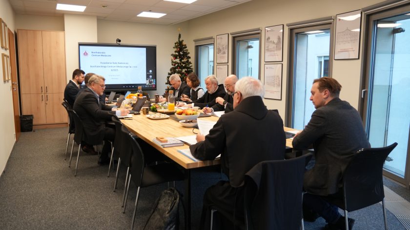 Członkowie Rady Nadzorczej Bonifraterskiego Centrum Medycznego siedzą przy długim stole konferencyjnym, pracując na laptopach i robiąc notatki. Na ekranie w tle widać prezentację dotyczącą posiedzenia. Na stole stoją owoce, napoje i dokumenty