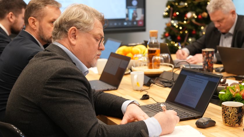 Mężczyzna w okularach i ciemnej marynarce siedzi przy stole konferencyjnym, robiąc odręczne notatki. Obok niego znajdują się laptopy, kubki oraz poczęstunek. W tle widać pozostałych uczestników posiedzenia.