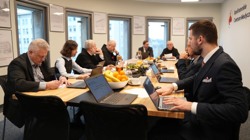 Uczestnicy spotkania siedzą wzdłuż stołu konferencyjnego, pracując na laptopach i zapisując notatki. Na stole ustawione są owoce, napoje i dokumenty. W tle widoczne są okna oraz plakaty z rysunkami obiektów Bonifraterskich