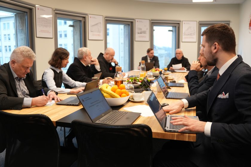 Uczestnicy spotkania siedzą wzdłuż stołu konferencyjnego, pracując na laptopach i zapisując notatki. Na stole ustawione są owoce, napoje i dokumenty. W tle widoczne są okna oraz plakaty z rysunkami obiektów Bonifraterskich
