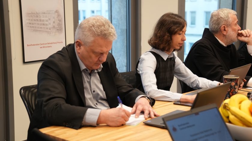 Mężczyzna w ciemnej marynarce zapisuje coś na kartce, a obok niego kobieta w białej koszuli i ciemnej sukience pracuje przy laptopie. W tle siedzą bracia zakonni biorący udział w posiedzeniu