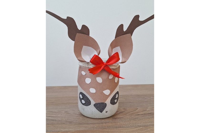 Ozdobny słoik przedstawiający renifera, z papierowymi rogami i czerwoną kokardką, ustawiony na drewnianym blacie