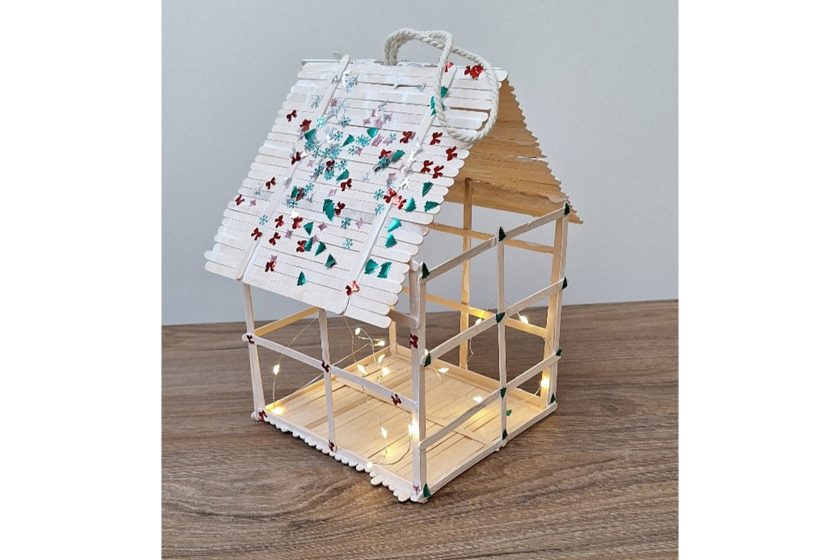 Domek-lampion wykonany z drewnianych patyczków, z podświetleniem LED i zdobionym dachem, stojący na drewnianym blacie.