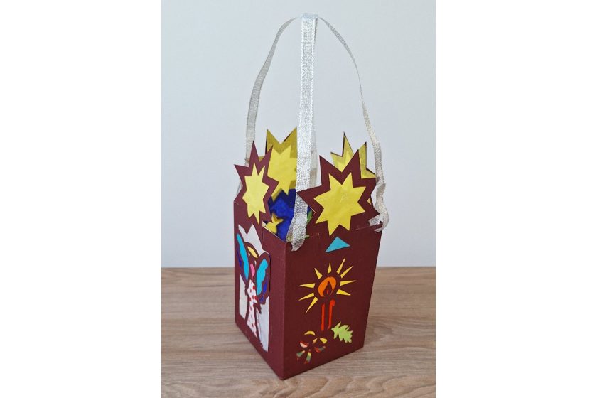 Czerwony kartonowy lampion z uchwytem z jasnej wstążki, ozdobiony gwiazdami, świecą i kolorowymi wycięciami