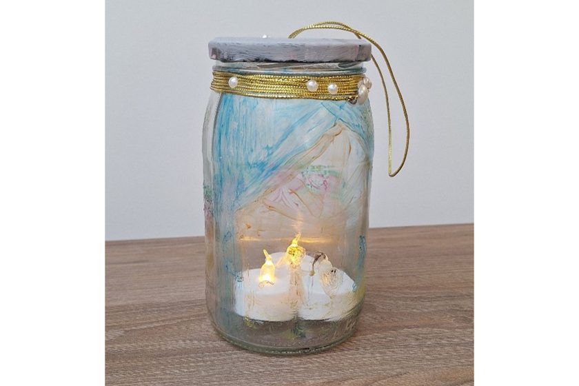 Szklany słoik ozdobiony malowanymi wzorami i złotym sznurkiem z perełkami, wewnątrz świeczka LED dająca ciepłe światło, stojący na drewnianej powierzchni
