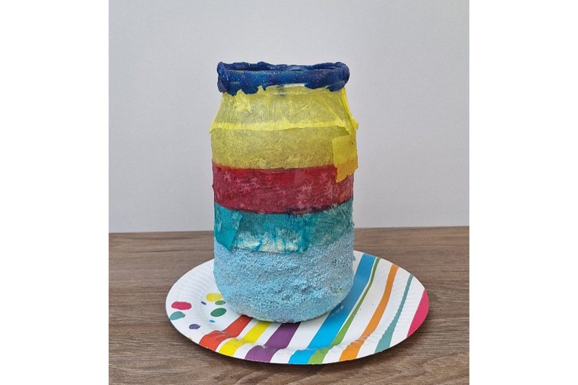 Szklany słoik ozdobiony warstwami kolorowej bibuły, ustawiony na papierowym talerzyku z wielobarwnym wzorem