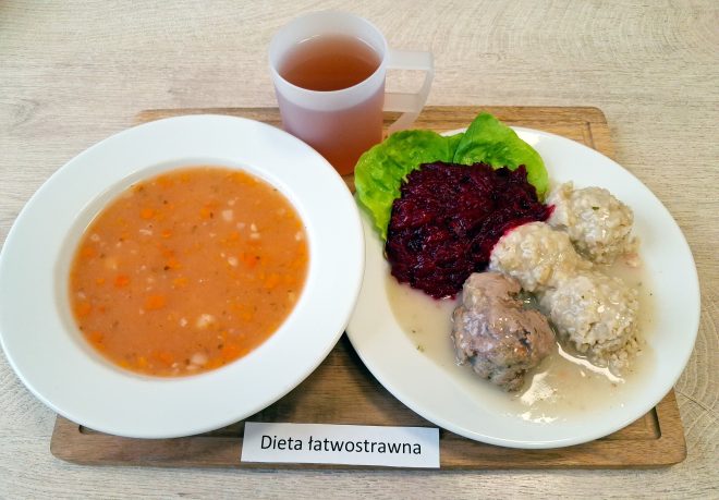 Talerz zupy jarzynowej o delikatnej, lekko pomarańczowej barwie, obok którego znajduje się drugie danie: pulpety w jasnym sosie, podane z buraczkami na liściu sałaty. Zestaw uzupełnia kubek kompotu. Całość ułożona na tacy z podpisem „Dieta łatwostrawna”