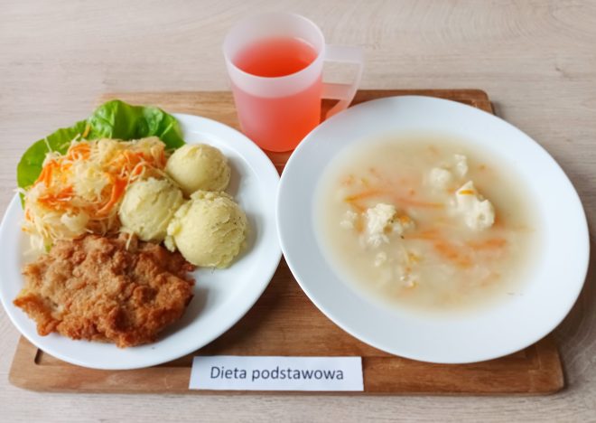 Na talerzu znajduje się obiad w diecie podstawowej: panierowany kotlet schabowy, trzy porcje puree ziemniaczanego oraz surówka z białej kapusty i marchewki na liściu sałaty. Obok podano talerz z lekką zupą warzywną oraz kubek czerwonego napoju. Zestaw opisany jest etykietą „Dieta podstawowa”