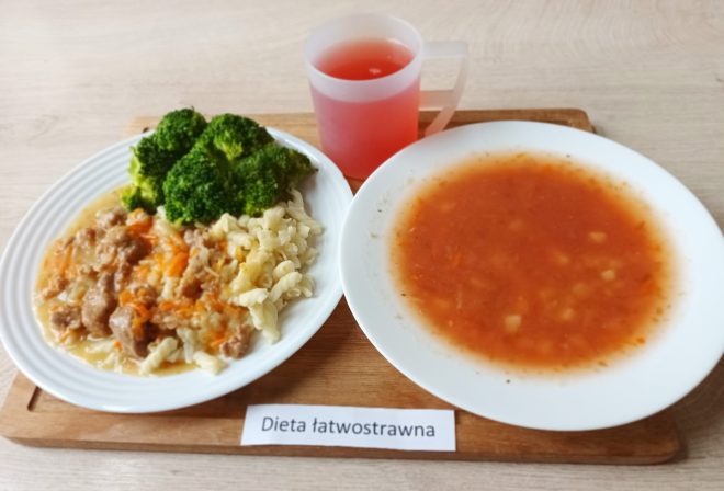 Obiad opisany jako Dieta łatwostrawna. Na białym talerzu znajduje się porcja duszonego mięsa z warzywami, ugotowany makaron typu świderki oraz gotowane różyczki brokuła. Obok ustawiono drugi talerz z jasnoczerwoną, warzywną zupą. Z tyłu stoi kubek z różowym napojem. Całość podana jest na drewnianej tacy z etykietą ‘Dieta łatwostrawna