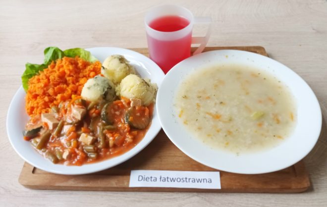 Na talerzu podano obiad w diecie łatwostrawnej: gotowane ziemniaki, duszone warzywa z sosem oraz porcję drobno startego gotowanego marchwiowego puree na liściu sałaty. Obok znajduje się talerz z lekką zupą warzywną oraz kubek czerwonego napoju. Zestaw oznaczony etykietą „Dieta łatwostrawna”