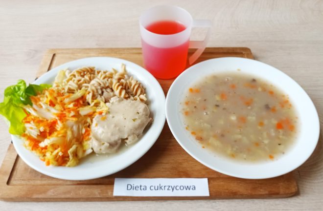 Talerz z pełnoziarnistym makaronem świderki, duszonym mięsem w sosie oraz surówką z marchwi i kapusty podaną na liściu sałaty. Obok zupa jarzynowa i kubek czerwonego kompotu. Zestaw oznaczony etykietą „Dieta cukrzycowa”