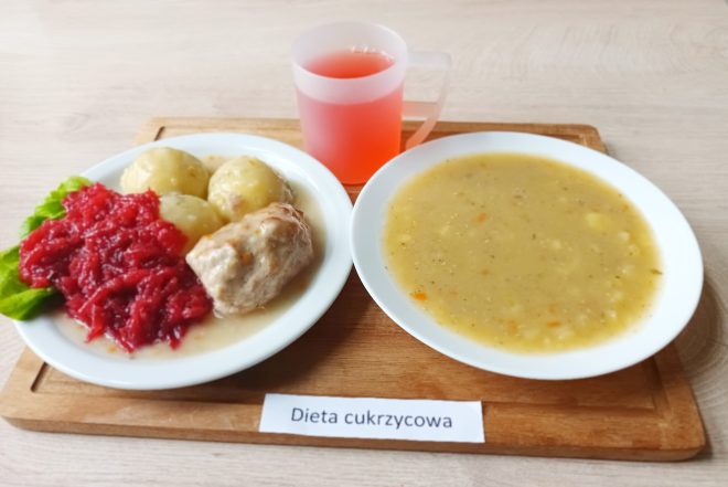 Talerz z gotowanymi ziemniakami, duszonym mięsem w jasnym sosie oraz porcją tartych buraczków na liściu sałaty. Obok miseczka zupy jarzynowej i kubek czerwonego kompotu. Zestaw ustawiony na tacy z etykietą „Dieta cukrzycowa”