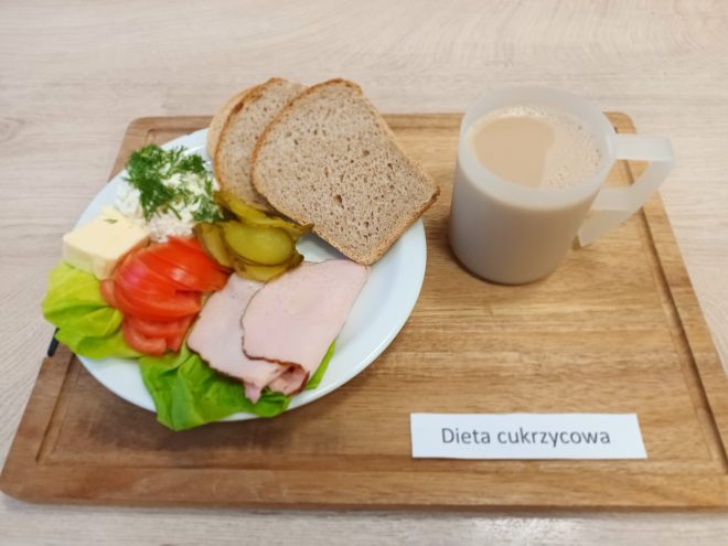 Na talerzu ułożono dwa plasterki wędliny, kilka plasterków pomidora, plasterki ogórka kiszonego, porcję białego sera z koperkiem, masło oraz dwie kromki pełnoziarnistego pieczywa. Obok stoi kubek kawy z mlekiem. Na tacy widoczna jest etykieta „Dieta cukrzycowa”