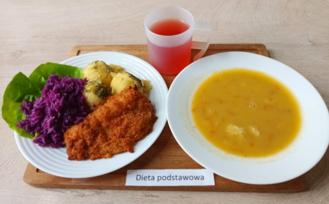 Obiad w diecie podstawowej: panierowany kotlet, ziemniaki z ziołami, surówka z czerwonej kapusty i liścia sałaty, talerz zupy jarzynowej oraz napój owocowy, podane na drewnianej desce