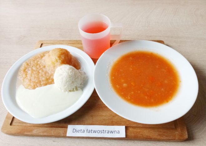 Obiad w diecie łatwostrawnej: talerz z zupą jarzynową o gładkiej konsystencji oraz drugie danie z gotowanym ryżem, mięsem drobiowym i sosem. Obok kubek z napojem owocowym. Posiłek podany na drewnianej tacy z etykietą „Dieta łatwostrawna”