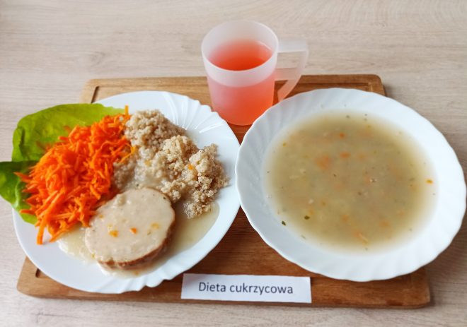 Obiad w diecie cukrzycowej: talerz z porcją mięsa w jasnym sosie, kaszą, surówką z tartej marchewki na liściu sałaty oraz miską zupy jarzynowej; obok kubek z napojem