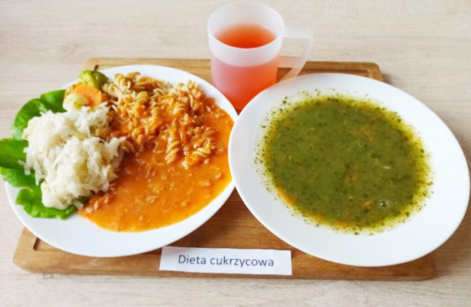 Porcja obiadowa w diecie cukrzycowej: talerz z zupą jarzynową o zielonym kolorze oraz drugie danie z makaronem w sosie pomidorowym, surówką z białej kapusty i liści sałaty. Obok kubek z napojem. Posiłek podany na drewnianej desce z etykietą „Dieta cukrzycowa”