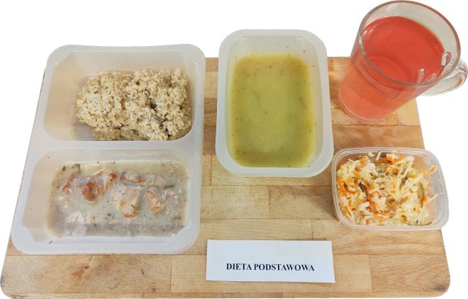 Na zdjęciu widać obiad w diecie podstawowej podany na plastikowej tacce: porcję kaszy, danie mięsne w sosie oraz pojemnik z zupą krem. Obok znajdują się małe pudełko z surówką z kapusty i marchewki oraz szklanka różowego kompotu. Zestaw ułożony jest na drewnianej desce, przed którą znajduje się karteczka z napisem „Dieta podstawowa”
