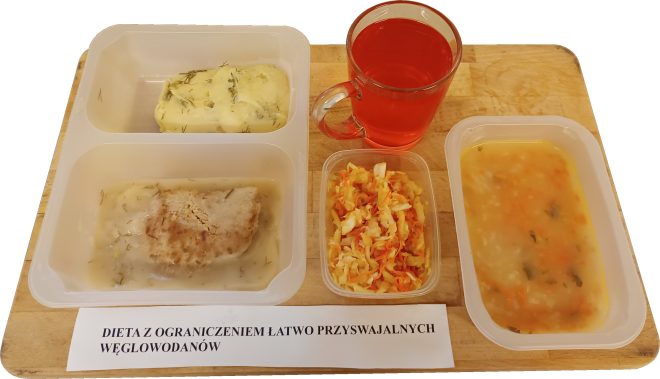 Na drewnianej tacy stoi zestaw szpitalnego obiadu w pojemnikach jednorazowych. W lewym górnym pojemniku znajduje się porcja puree ziemniaczanego posypanego koperkiem. W lewym dolnym pojemniku leży kotlet mielony polany jasnym sosem. Obok, w mniejszym pojemniczku, znajduje się surówka z białej kapusty i marchwi. Po prawej stronie ustawiono pojemnik z zupą jarzynową o żółtawym kolorze. Do posiłku dołączono szklankę czerwonego kompotu. Przed tacą leży karteczka z napisem „Dieta z ograniczeniem łatwo przyswajalnych węglowodanów”