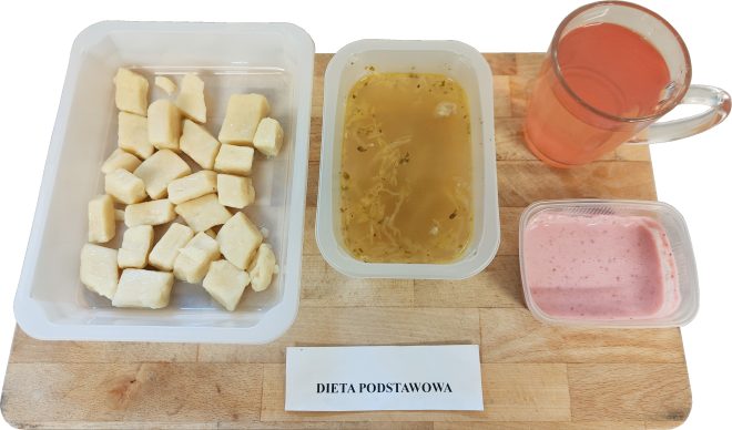 Obiad w diecie podstawowej: w pojemniku kluski kładzione, obok pojemnik z jarzynową zupą o klarownej bazie, różowy deser mleczny oraz kubek kompotu. Zestaw ustawiony na desce z podpisem ‘Dieta podstawowa