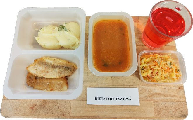 Zestaw obiadowy w pojemnikach jednorazowych opisany jako Dieta Podstawowa. W lewym pojemniku z trzema przegródkami znajduje się porcja puree ziemniaczanego z koperkiem oraz dwa kawałki panierowanej ryby. Obok stoi prostokątny pojemnik z zupą o pomarańczowym kolorze, najprawdopodobniej pomidorową lub jarzynową. Po prawej stronie umieszczono kubek z czerwonym napojem oraz mały pojemnik z surówką z białej kapusty, marchwi i ogórka