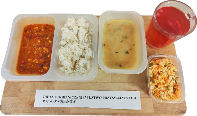 Obiad w diecie z ograniczeniem łatwo przyswajalnych węglowodanów podany na drewnianej desce. Na plastikowej tacce znajdują się trzy porcje: gęsty sos pomidorowy z warzywami i mięsem, biała gotowana ryżowa kasza oraz jasna zupa z warzywami i ziołami. Obok tacki stoi małe pudełko z surówką z kapusty, marchwi i ogórka oraz szklanka czerwonego kompotu. Przed zestawem leży karteczka z nazwą diety