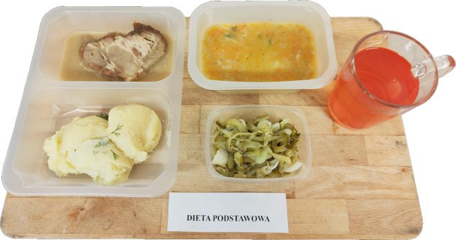 Zestaw obiadowy w pojemnikach: gotowane mięso w sosie, ziemniaki puree z koperkiem, surówka z kiszonej kapusty oraz miseczka zupy jarzynowej. Obok szklanka czerwonego kompotu. Z przodu widoczna etykieta z napisem „Dieta podstawowa”