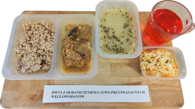 Na tacy znajduje się obiad w diecie z ograniczeniem łatwo przyswajalnych węglowodanów: porcja kaszy pęczak, mięso w sosie ułożone w osobnej przegródce, zupa jarzynowa w pojemniku oraz surówka z białej kapusty i marchwi. Obok stoi przezroczysty kubek z czerwonym kompotem