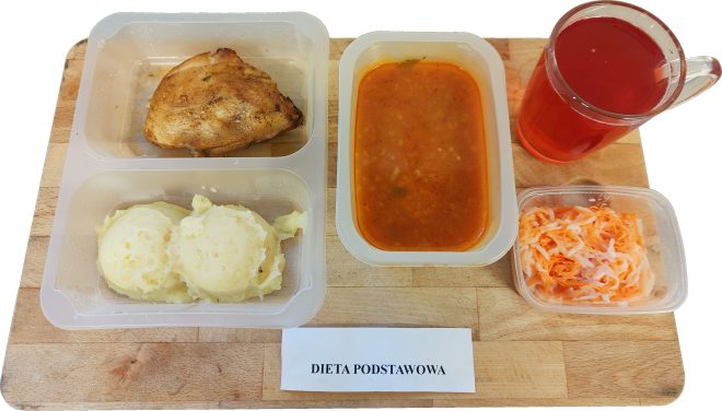 Obiad w diecie podstawowej składa się z porcji pieczonego udka z kurczaka, ziemniaków puree oraz gęstej zupy pomidorowej w osobnym pojemniku. Obok znajduje się surówka z marchwi i białej rzodkwi oraz kubek czerwonego napoju. Pod tacą widnieje etykieta „Dieta podstawowa”