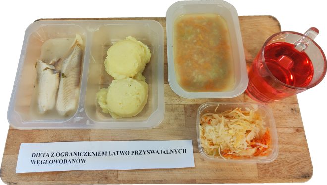 Posiłek szpitalny w diecie z ograniczeniem łatwo przyswajalnych węglowodanów: filet rybny w jasnym sosie, porcja puree ziemniaczanego, zupa jarzynowa, surówka z kiszonej kapusty oraz napój owocowy, podane na drewnianej desce