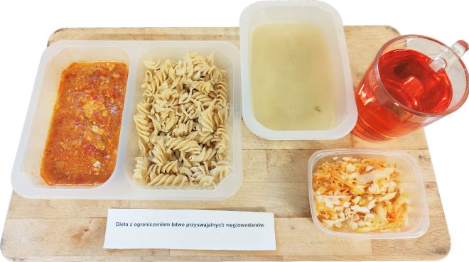 Obiad w diecie z ograniczeniem łatwo przyswajalnych węglowodanów: makaron pełnoziarnisty z sosem pomidorowym, porcja klarownej zupy, surówka z kapusty i marchwi oraz herbata, podane na drewnianej tacy
