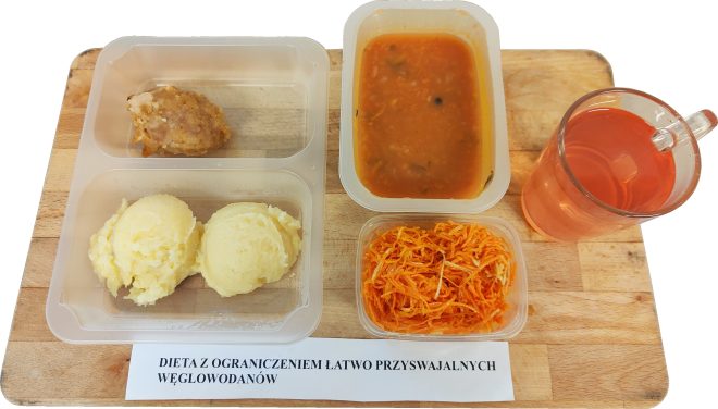Obiad w diecie z ograniczeniem łatwo przyswajalnych węglowodanów: kotlet mielony, puree ziemniaczane, surówka z marchewki, zupa pomidorowa oraz kompot, podane w pojemnikach na drewnianej desce