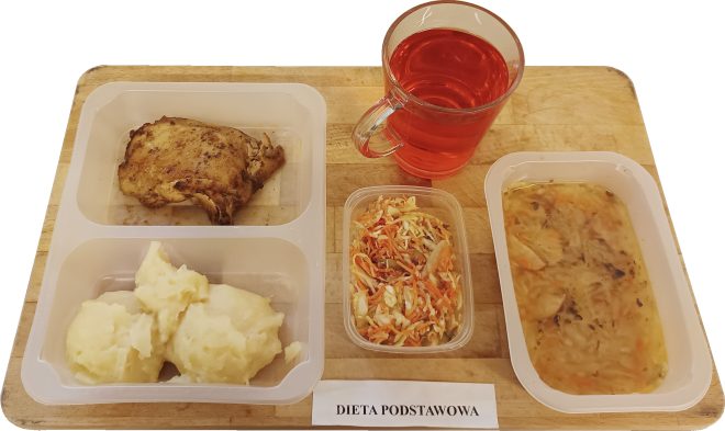 Obiad w diecie podstawowej: pieczone udko z kurczaka, ziemniaki puree, surówka z kapusty i marchewki, zupa kapuśniak oraz kompot, podane na tacy na drewnianej desce
