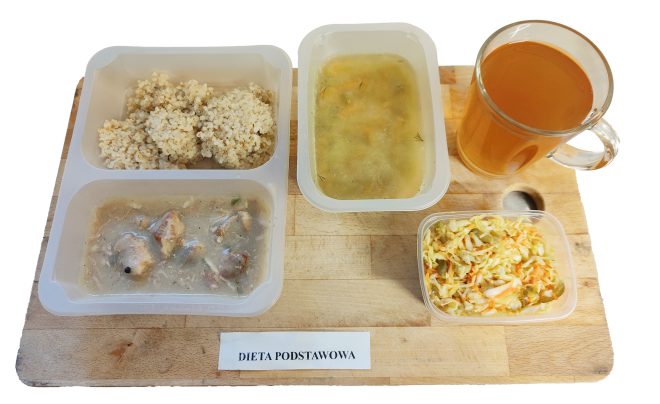 Zestaw obiadowy w diecie podstawowej: zupa jarzynowa, kasza jęczmienna, mięso w sosie oraz surówka z kapusty, podane w pojemnikach na drewnianej desce. Obok kubek herbaty