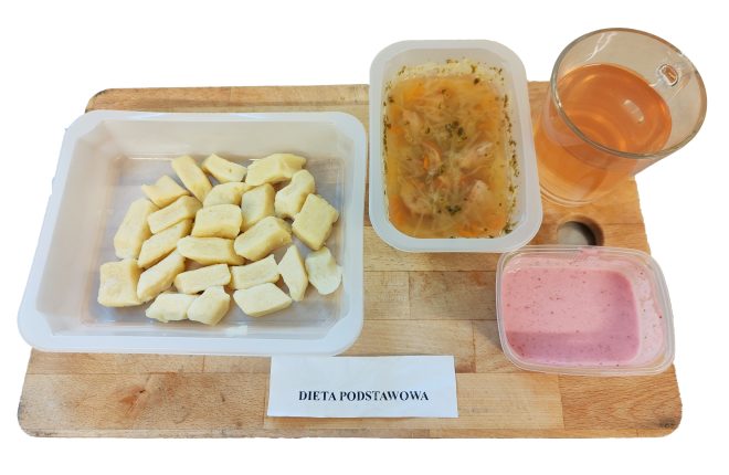 Obiad w diecie podstawowej: kluski kładzione podane w pojemniku, obok porcja zupy jarzynowej z makaronem, deser w postaci różowego budyniu lub musu oraz herbata. Posiłki ułożone na drewnianej desce, z podpisem „Dieta podstawowa”