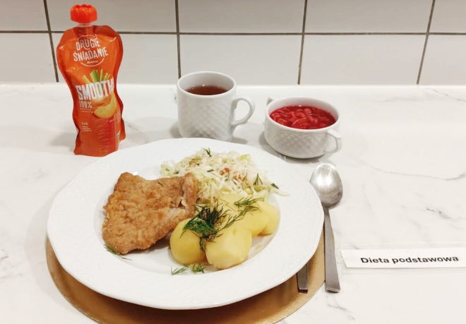 Talerz z panierowanym kotletem, gotowanymi ziemniakami z koperkiem i surówką z białej kapusty. Obok filiżanka herbaty, miseczka czerwonego barszczu i saszetka smoothie. Podpis: Dieta podstawowa.
