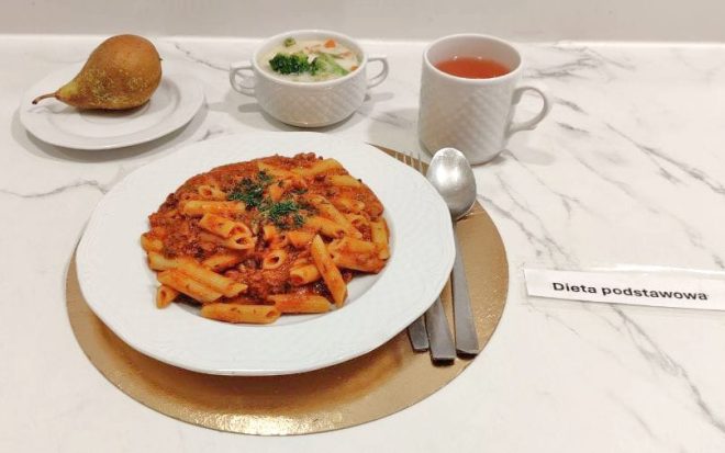 Na talerzu znajduje się porcja makaronu penne z sosem bolońskim, udekorowana ziołami. Obok ustawiono miseczkę z zupą jarzynową, kubek z herbatą oraz osobny talerzyk z gruszką. Zestaw oznaczony jest etykietą „Dieta podstawowa”