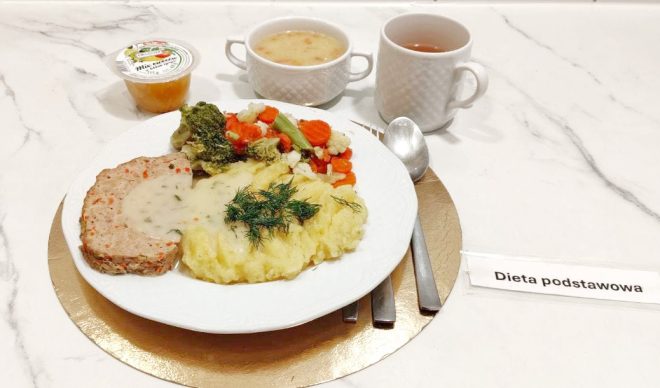 Talerz z obiadem zawiera pulpet w sosie, porcję puree ziemniaczanego z koperkiem oraz gotowane warzywa: brokuł, marchew, kalafior i fasolkę. W tle widoczne są miseczka z zupą, kubek z herbatą oraz deser w plastikowym kubeczku. Zestaw oznaczono etykietą „Dieta podstawowa”