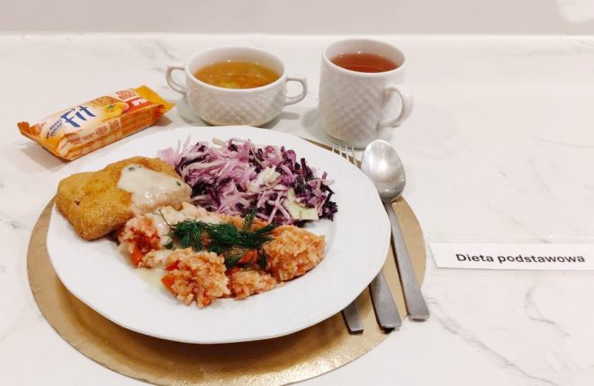 Obiad w diecie podstawowej podany na białym talerzu ustawionym na złotej podkładce. Na talerzu znajdują się: panierowany kotlet polany jasnym sosem, porcja kaszy lub ryżu z warzywami udekorowana koperkiem oraz surówka z białej i czerwonej kapusty. W tle stoją dwie białe filiżanki — jedna z przezroczystym bulionem, druga z herbatą — a obok leży baton zbożowy. Po prawej stronie widoczna jest karteczka z napisem „Dieta podstawowa”