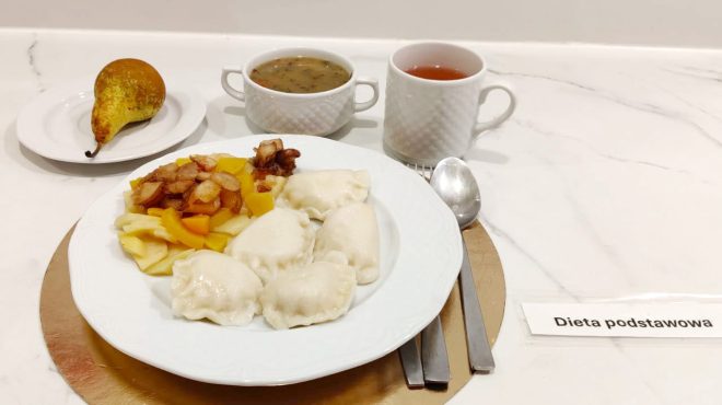 Talerz z porcją pierogów oraz gotowanymi warzywami podsmażonymi z cebulką. W tle miseczka zupy, kubek herbaty oraz talerzyk z gruszką. Przy talerzu leżą sztućce, a obok znajduje się etykieta „Dieta podstawowa”