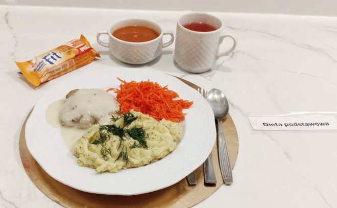 Obiad w diecie podstawowej: pulpety w jasnym sosie, ziemniaki z koperkiem oraz surówka z tartej marchewki; w tle miska zupy i kubek herbaty, całość podana na jasnym blacie