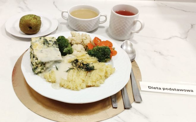 Obiad w diecie podstawowej: zapiekanka warzywna ze szpinakiem podana z ziemniakami puree i gotowanymi warzywami. Obok filiżanki zupy, herbaty oraz jabłko na deser