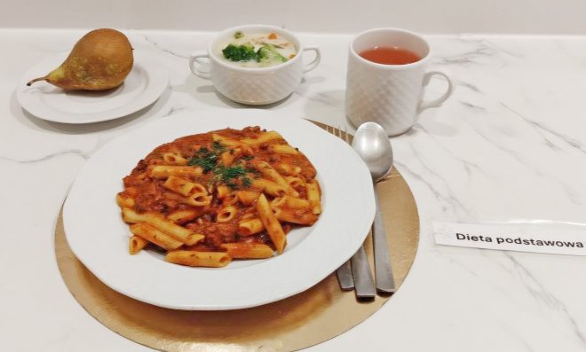 Talerz makaronu penne z sosem pomidorowym i ziołami, obok miseczka z zupą warzywną i kubek herbaty. Z lewej strony na talerzyku leży gruszka. Zestaw opisany jako dieta podstawowa