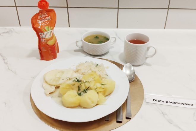 Talerz obiadowy z gotowanymi ziemniakami posypanymi koperkiem, gotowaną piersią kurczaka w sosie oraz porcją białej surówki. W tle miseczka zupy jarzynowej, kubek herbaty oraz saszetka owocowego smoothie. Obok talerza leżą sztućce i etykieta „Dieta podstawowa”