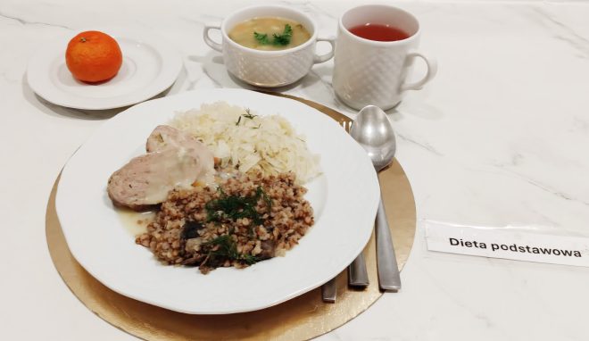 Obiad w diecie podstawowej: plasterki pieczonego mięsa w sosie, kasza gryczana oraz duszona kapusta. W tle miseczka zupy, herbata i mandarynka na talerzyku