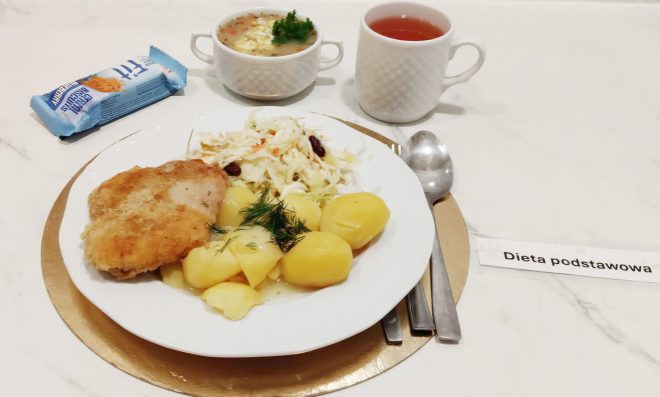 Obiad w diecie podstawowej: panierowany kotlet drobiowy, gotowane ziemniaki z koperkiem oraz surówka z białej kapusty. Na stole miseczka zupy i kubek herbaty