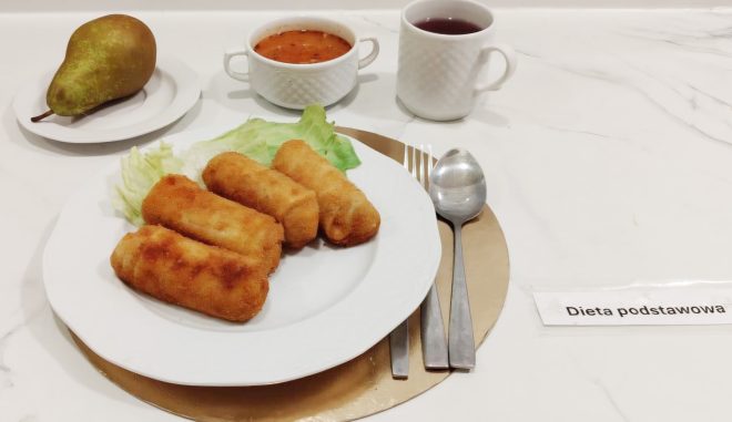 Obiad w diecie podstawowej: cztery krokiety na liściach sałaty, zupa w białej filiżance, herbata oraz gruszka na małym talerzu