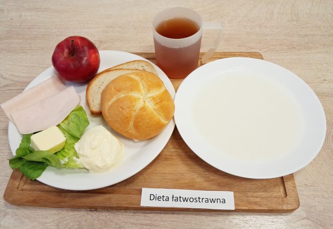 Zestaw śniadaniowy: bułka i kromki białego pieczywa, plasterki wędliny, masło, sałata, jabłko oraz kubek herbaty. Obok talerz z jasną zupą mleczną. Podpis: Dieta łatwostrawna